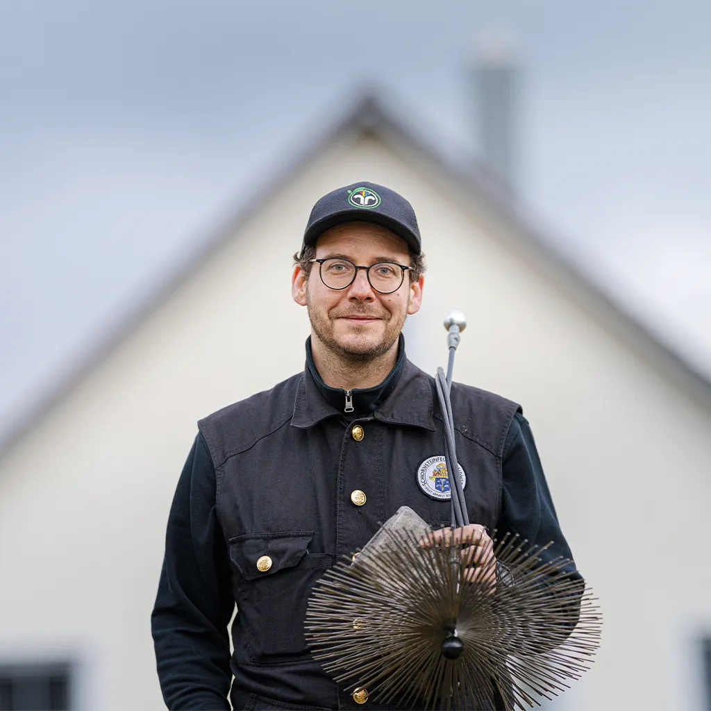 Oliver Heiden steht in Kaminkehrerkleidung mit Kaminkehrerwerkzeug vor einem Haus, blick nach vorne und lächelt.